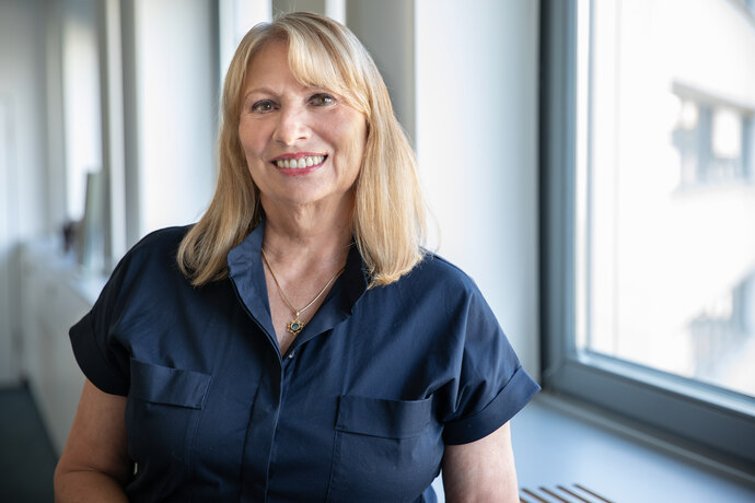 Porträtfoto einer lächelnden Frau mit schulterlangem blondem Haar und dunkelblauer Bluse; sie trägt eine Halskette. Aufnahme in einem hellen Flur mit großen Fenstern und unscharfem Hintergrund.